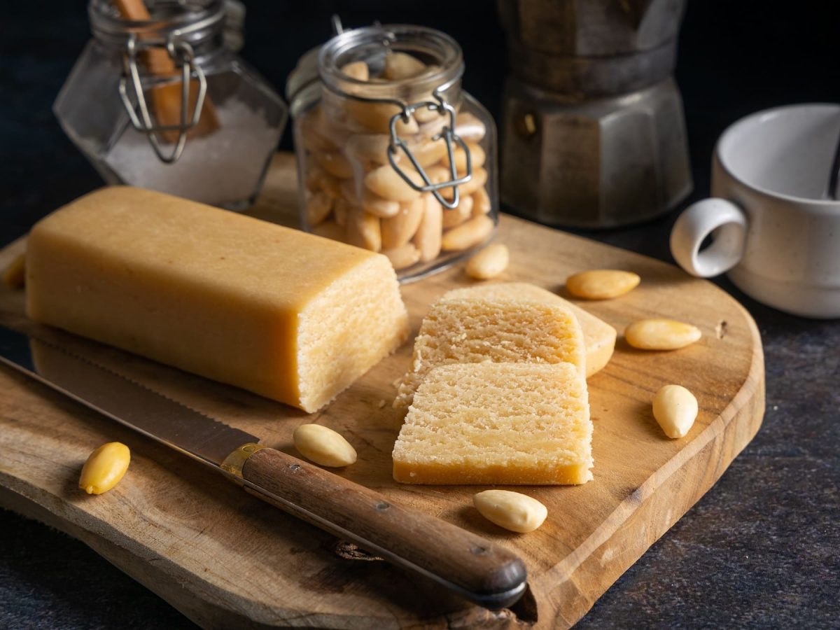 Ein Holzbrett mit Marzipan und dunklem Hintergrund. Darauf auch Mandeln, Zucker und Kaffee. Alles von der Seite fotografiert.