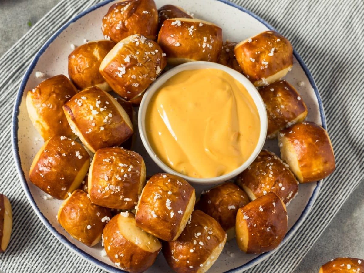 Brezel-Bites auf einem weißen Teller mit blauen Rand, in der Mitte ein Schüsselchen mit Käsesauce. Der Teller steht auf einem grau-weiß-gestreiften Geschirrtuch, der Hintergrund ist grau.