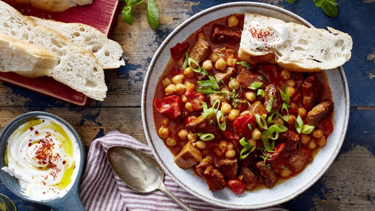 Orientalisches Rindsgulasch mit Weißbrot im Teller auf Holzuntergrund, drum herum Brotscheiben, Schale mit Joghurt, Teller und Küchentuch.