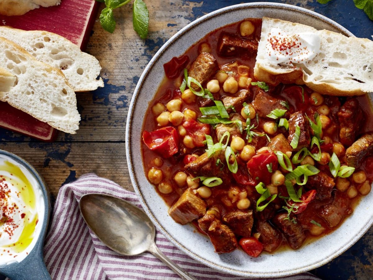 Orientalisches Rindsgulasch mit Weißbrot im Teller auf Holzuntergrund, drum herum Brotscheiben, Schale mit Joghurt, Teller und Küchentuch.