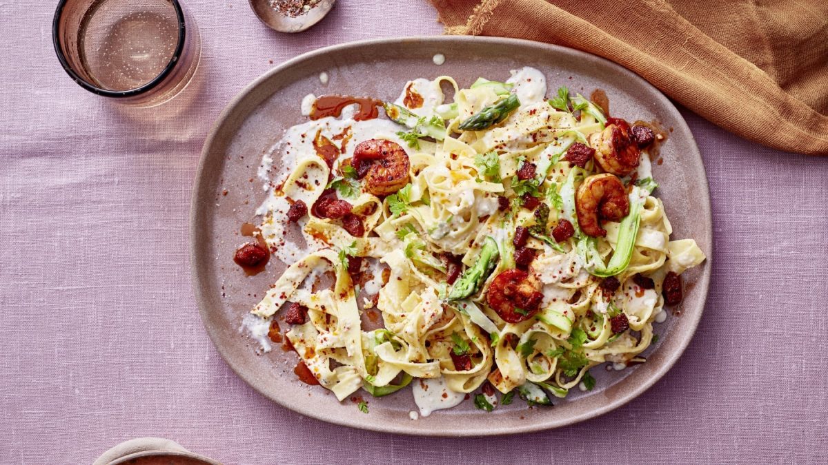 Pasta in Mandel-Lemon-Cream mit Garnelen und Chorizo auf rosa Teller mit gleichem Untergrund, drum herum Gläser und Küchentuch in ockergelb.