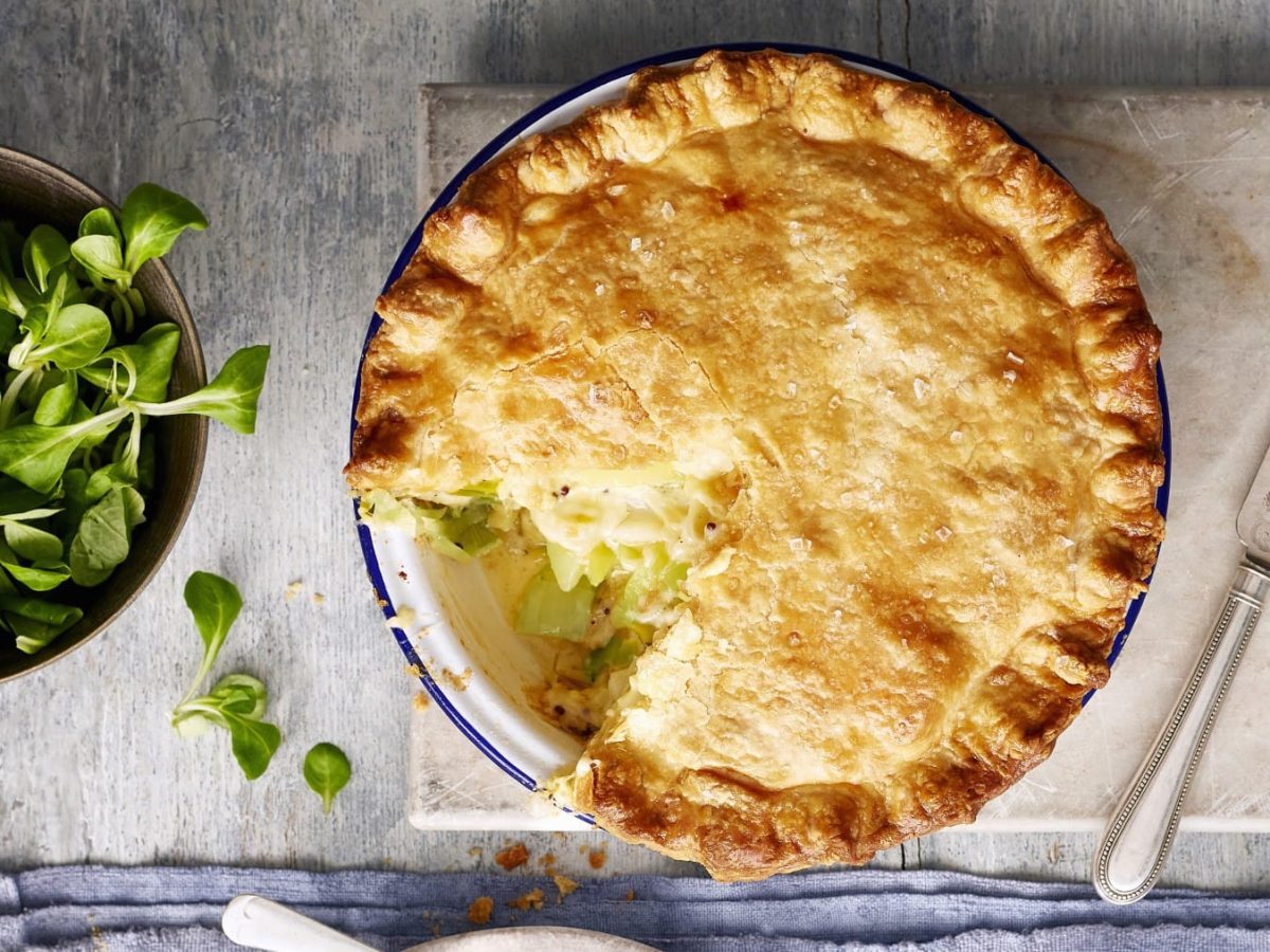 Angeschnittener Pie mit Kartoffel-Lauch-Füllung auf grauem Untergrund, daneben Schüssel mit Feldsalat.