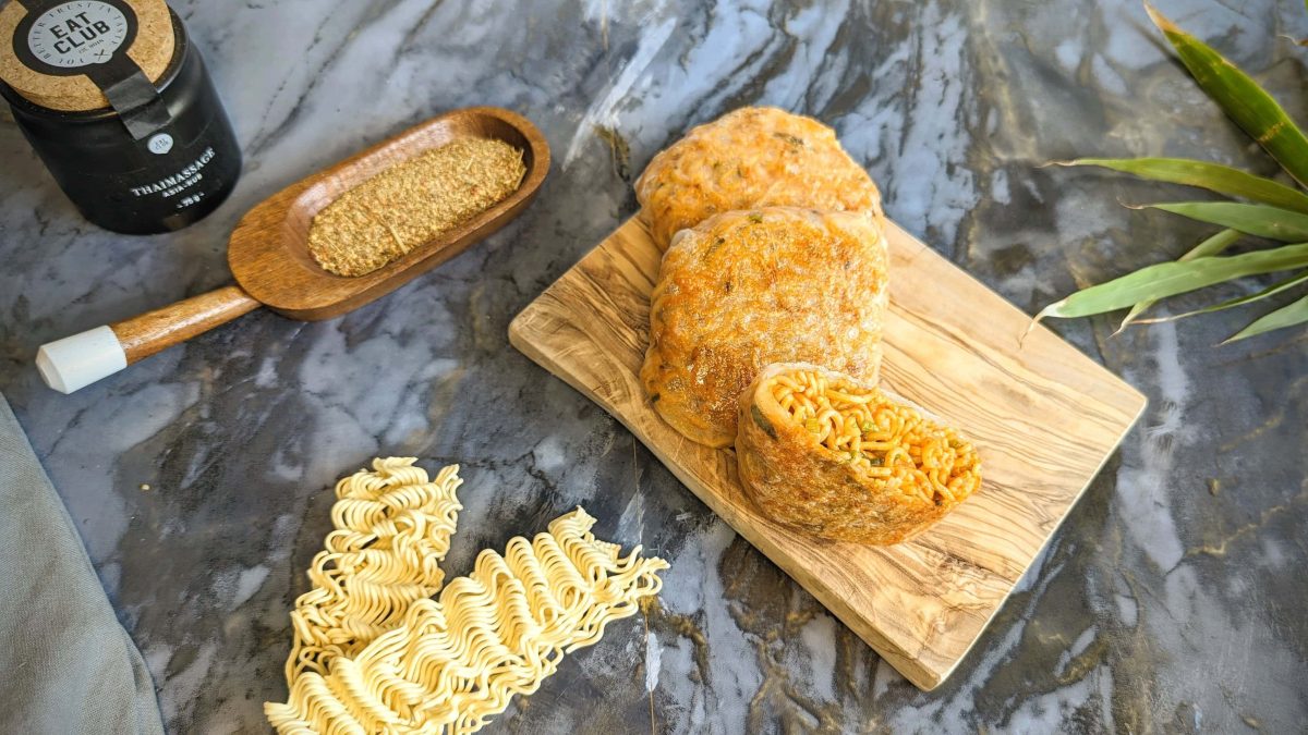 Zwei Hot Noodle Pockets auf einem kleinen Holzbrett udn grauem Steintisch. Daneben asiatische Nudeln, Sesam sowie asiatisches Gewürz. Alles von oben fotografiert.