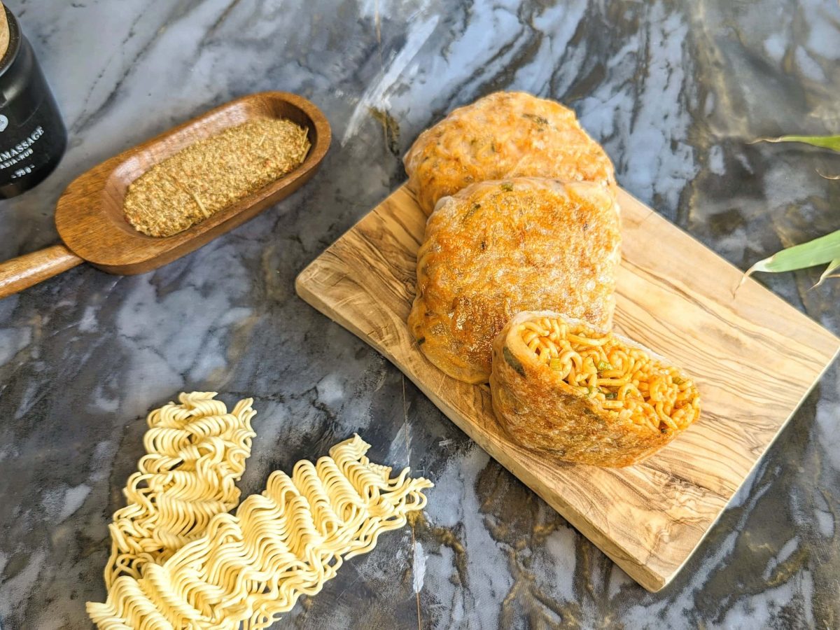 Zwei Hot Noodle Pockets auf einem kleinen Holzbrett udn grauem Steintisch. Daneben asiatische Nudeln, Sesam sowie asiatisches Gewürz. Alles von oben fotografiert.