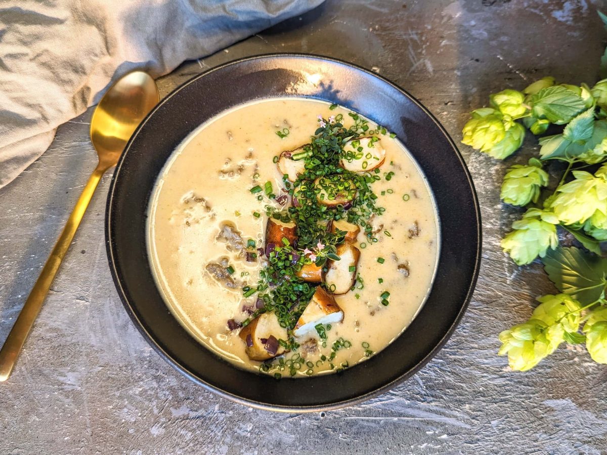 Eine schwarze Schüssel mit der Suppe auf einem grauen Untergrund. Daneben frischer Hopfen und ein goldener Löffel. Alles direkt von oben fotografiert.