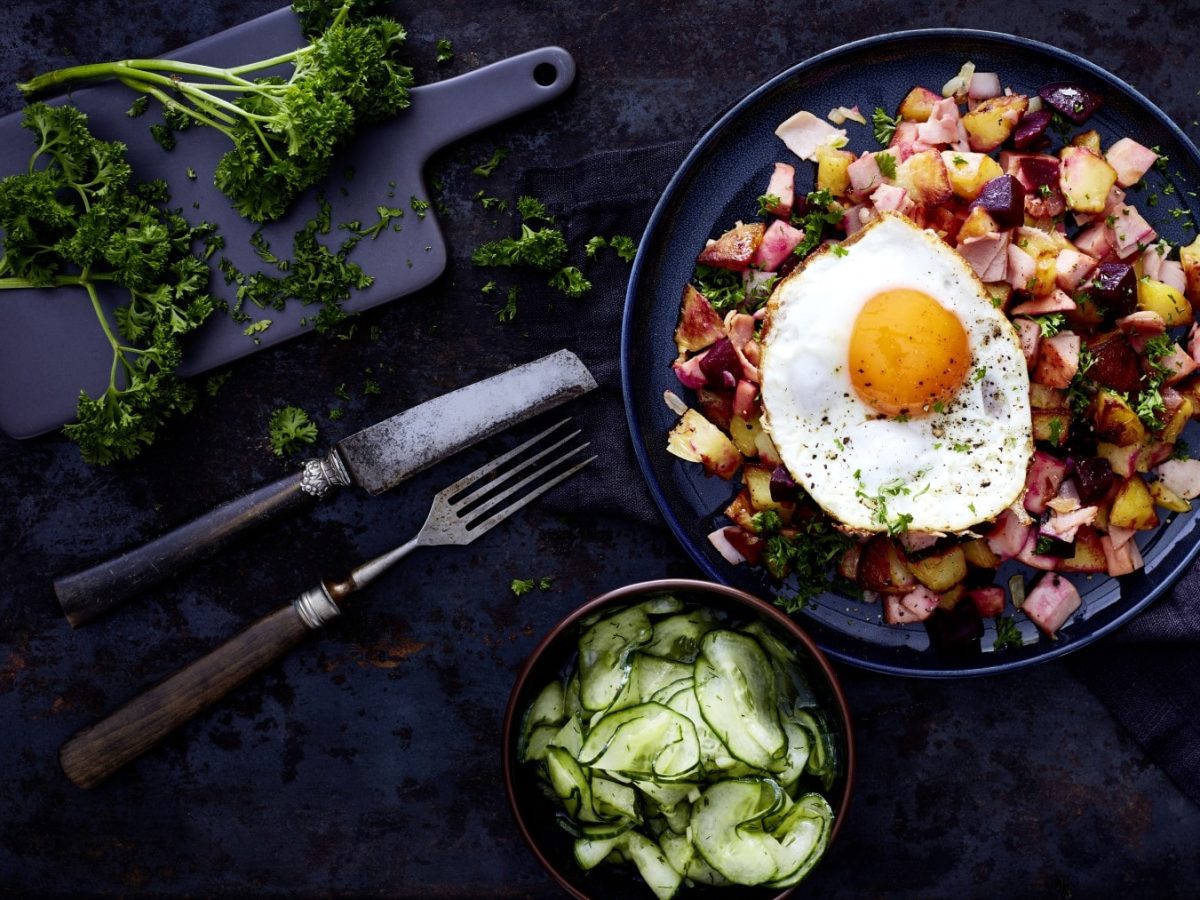 Pytt i Panna - schwedische Bratkartoffeln auf einem dunkelblauen Teller auf einem dunkelblauen Geschirrtuch auf einem dunkelblauen Hintergrund. Unten eine Schüssel mit Gurkensalat und Besteck. Links oben ein kleine Schneidebrett mit Petersilie.