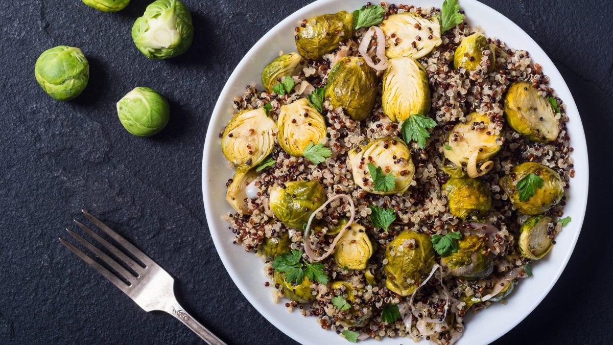 Ein weißer Teller mit dem Rosenkohl-Quinoa-Salat auf einem schwarzen Untergrund direkt von oben fotografiert. Auf der linken Seite noch frischer Rosenkohl und eine silberne Gabel.