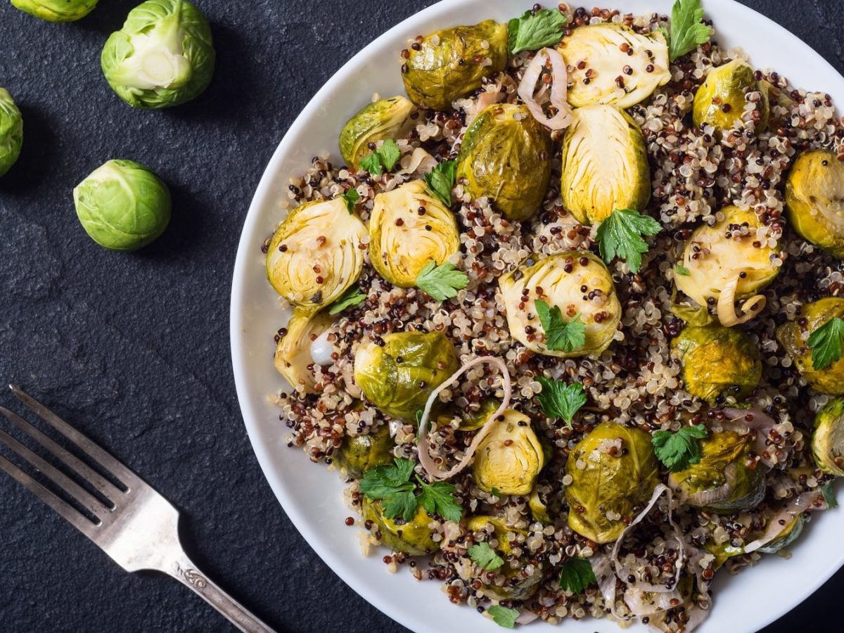 Ein weißer Teller mit dem Rosenkohl-Quinoa-Salat auf einem schwarzen Untergrund direkt von oben fotografiert. Auf der linken Seite noch frischer Rosenkohl und eine silberne Gabel.