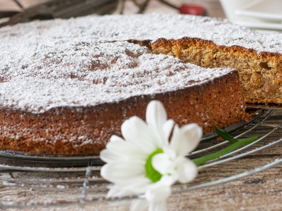 Ein angeschnittener Mandelkuchen auf einem Kuchengitter und hellem Holztisch sehr nahe von der Seite fotografiert.