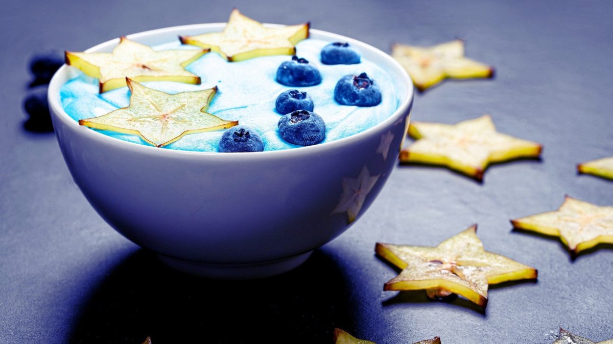 Frontal: eine weiße Schüssel mit einer Sternfrucht-Blaubeeren-Bowl auf blauem Untergrund. Drum herum liegen Scheiben einer Sternfrucht.