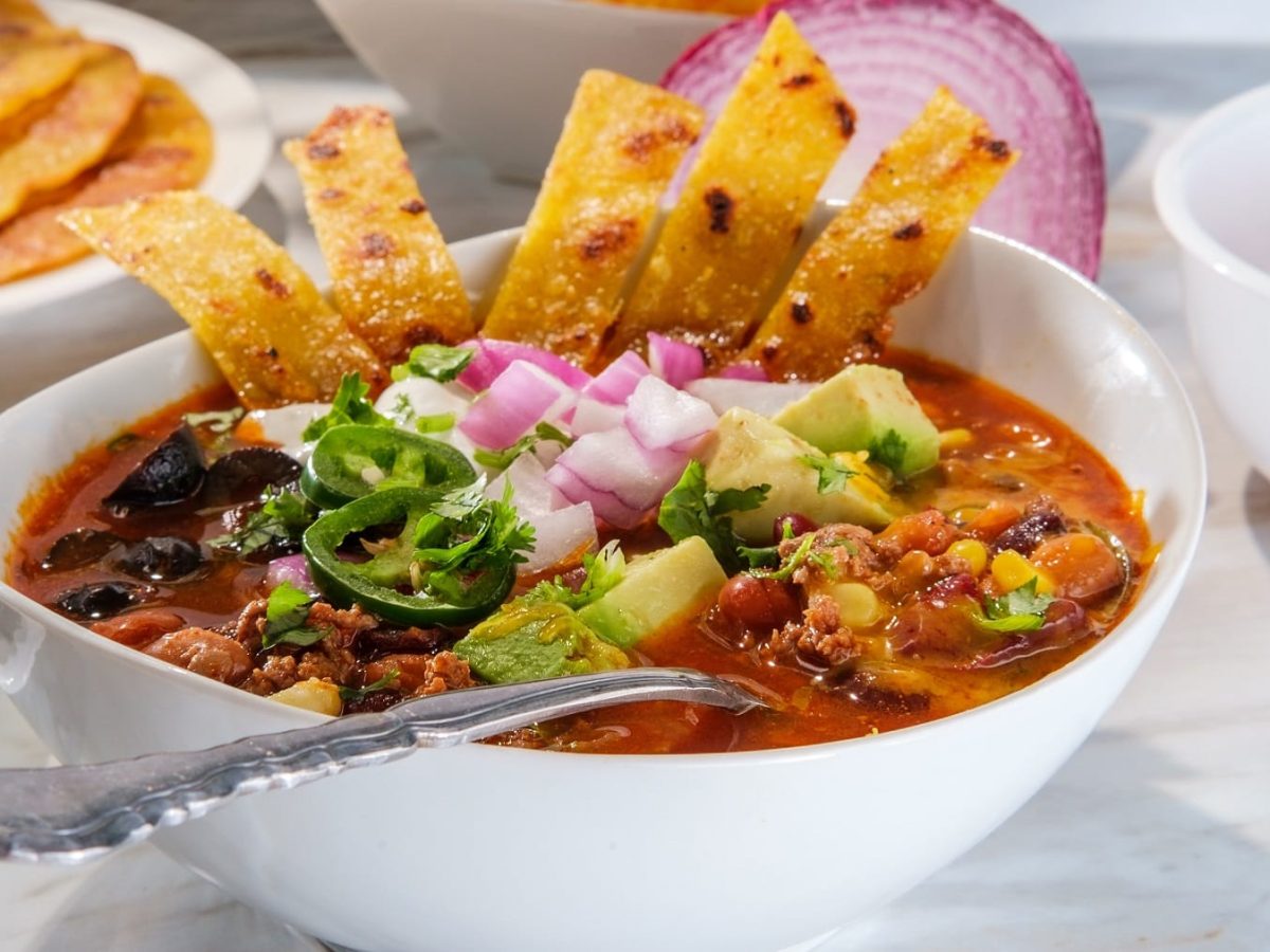 Eine weiße Schüssel mit der Taco-Suppe auf weißem Marmor. In der Suppe ein Löffel und etwas Mais-Fladenbrot. Alles von der Seite fotografiert.