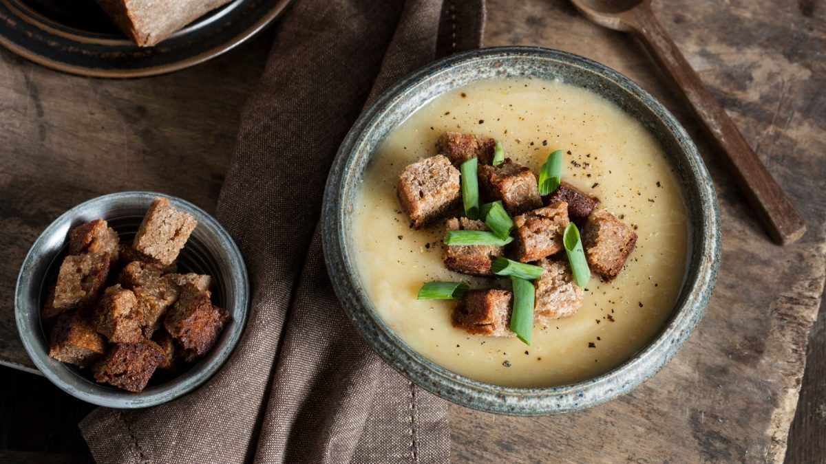 Eine Topinambur-Suppe mit knusprigen Brotwürfel auf dunkelbraunem Hintergrund. Links eine kleine Schüssel mit Brotwürfel, rechts oben ein Löffel aus Holz.