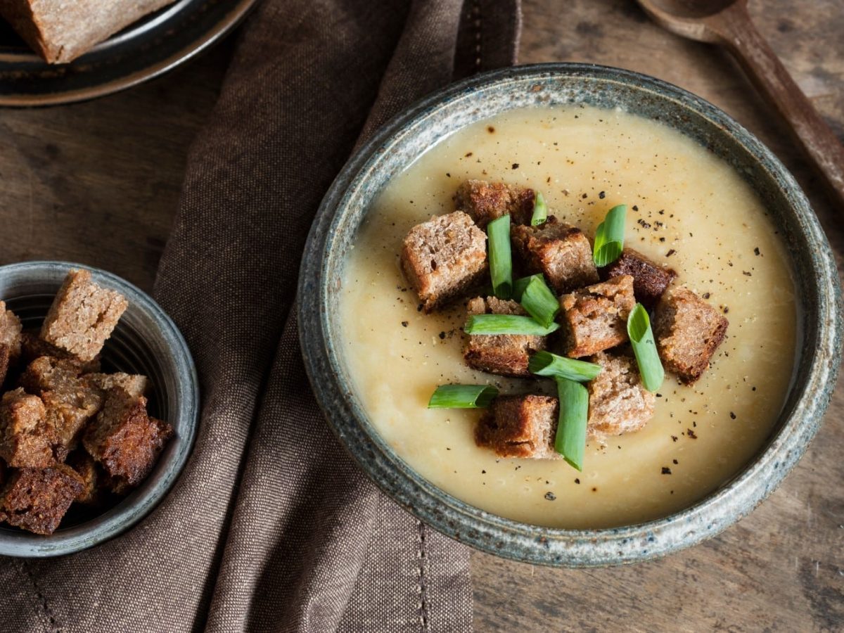 Eine Topinambur-Suppe mit knusprigen Brotwürfel auf dunkelbraunem Hintergrund. Links eine kleine Schüssel mit Brotwürfel, rechts oben ein Löffel aus Holz.