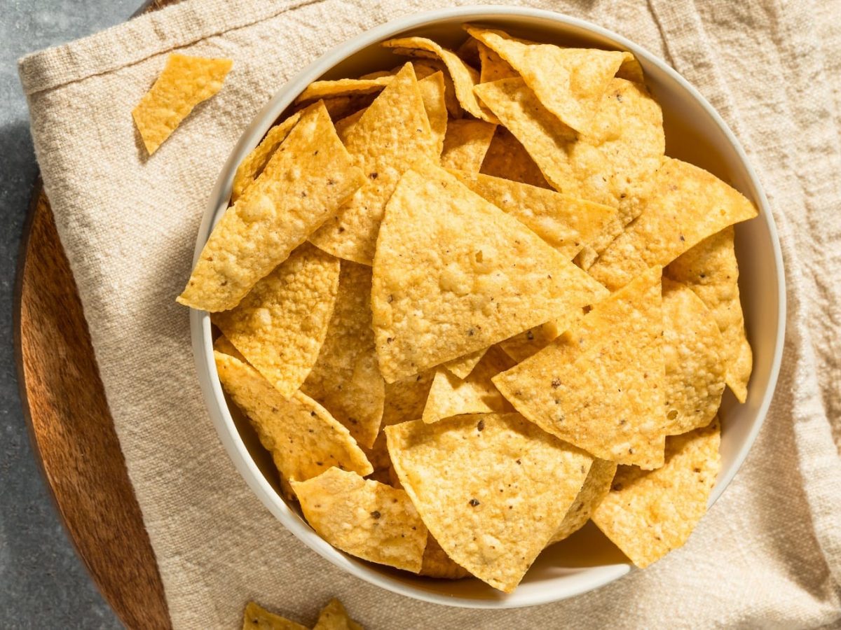 Eine weiße Schale mit den Tortilla Chips auf einer hellen Tischdecke und grauem Tisch. Alles direkt von oben fotografiert.