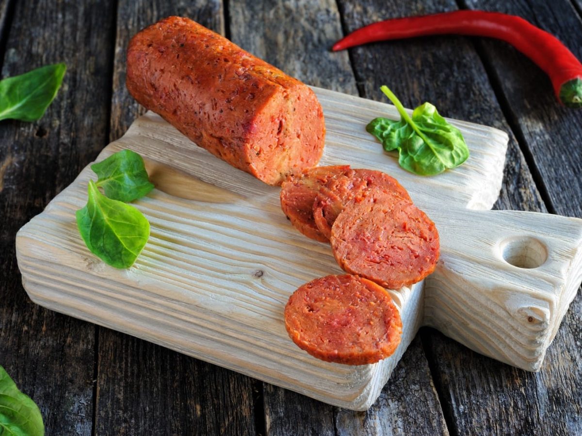 Vegane Salami auf einem Schneidebrett auf einem hölzernen Hintergrund. Oben auf dem Foto eine rote Chili. Auf dem Schneidebrett und dem Hintergrund Basilikumblätter verteilt.