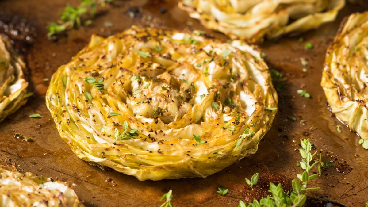 Gebackene Weißkohl-Steaks mit Feta-Creme von oben fotografiert. Alles auf einem dunklen Holzbrett mit Kräutern und grobem Pfeffer.