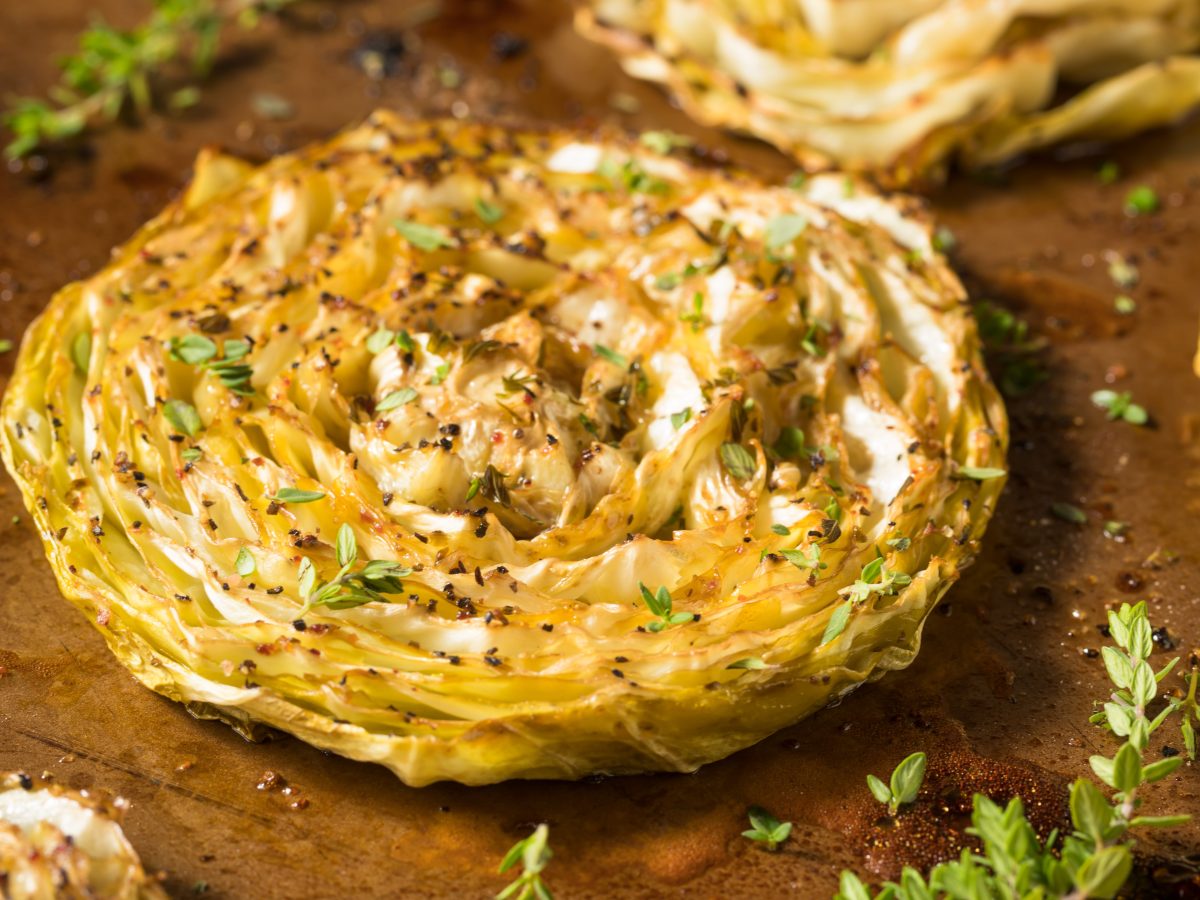 Gebackene Weißkohl-Steaks mit Feta-Creme von oben fotografiert. Alles auf einem dunklen Holzbrett mit Kräutern und grobem Pfeffer.