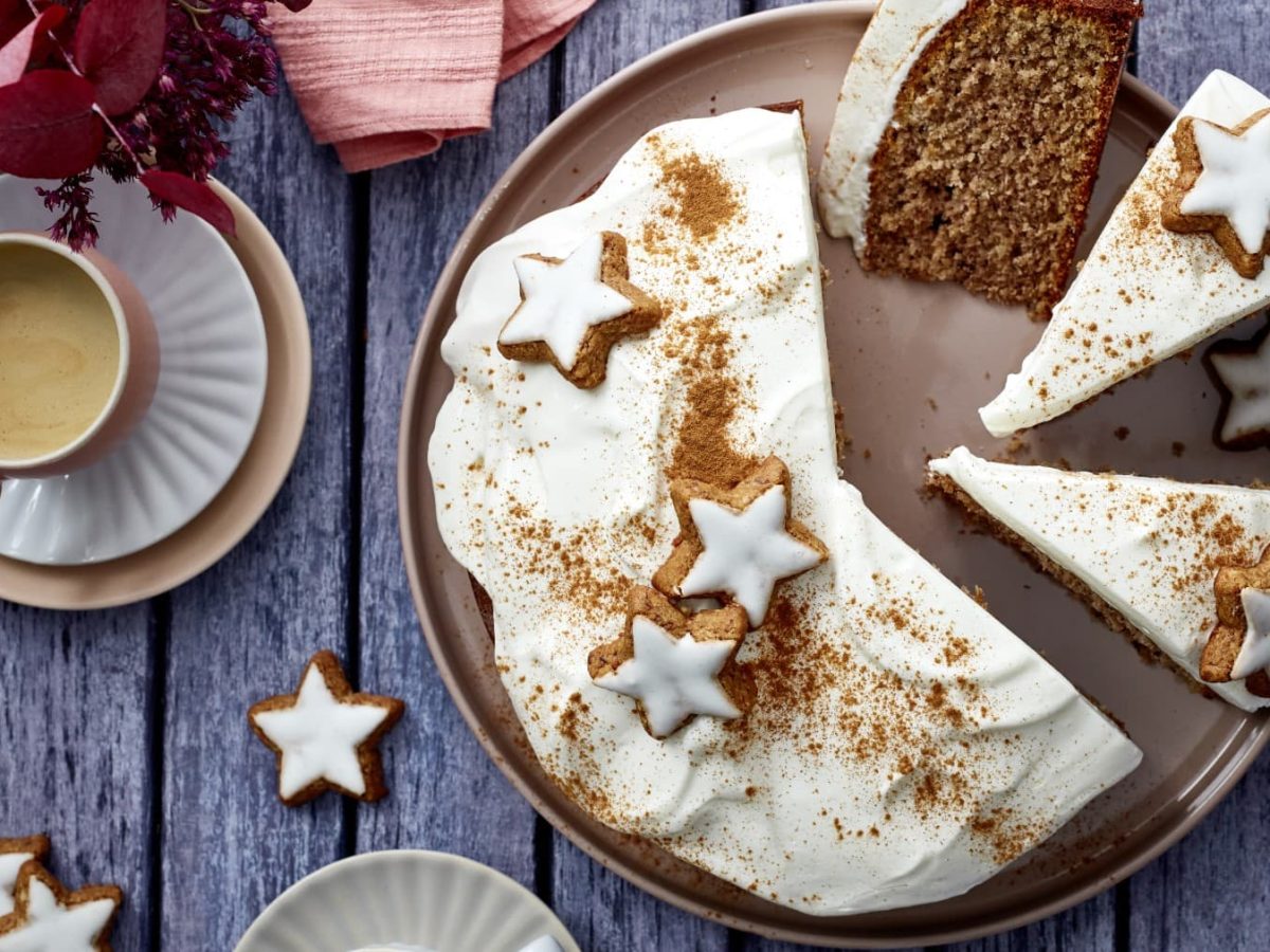 Ein Zimtstern-Nusskuchen auf einem braunen Teller auf einem dunkelgrauen Hintergrund. Links eine Tasse Kaffee und Blumen. Auf dem Hintergrund Zimtsterne verteilt.