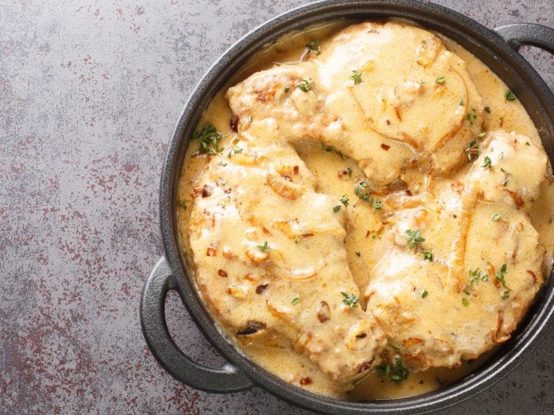 Foto von saftigen Zwiebel-Sahne-Schnitzeln, serviert in einer schwarzen, gusseisernen Pfanne. Die Schnitzel sind reichlich mit einer cremigen Zwiebelsauce bedeckt, die durch ihre glänzende Konsistenz und die sichtbaren Zwiebelstücke auffällt.