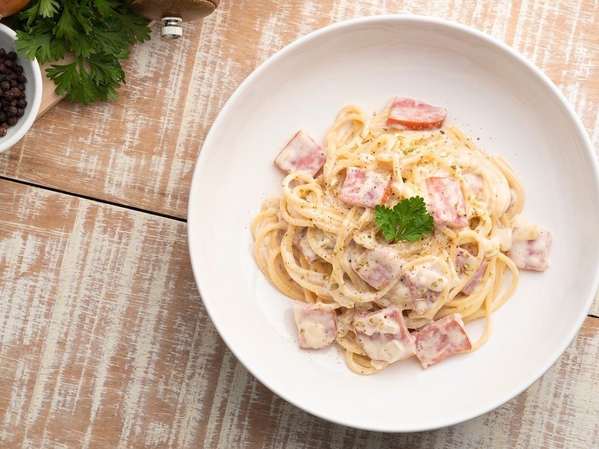 Ein weißer Teller mit Spaghetti mit Schinkensahnesauce auf einem hellbraunen Holztisch.