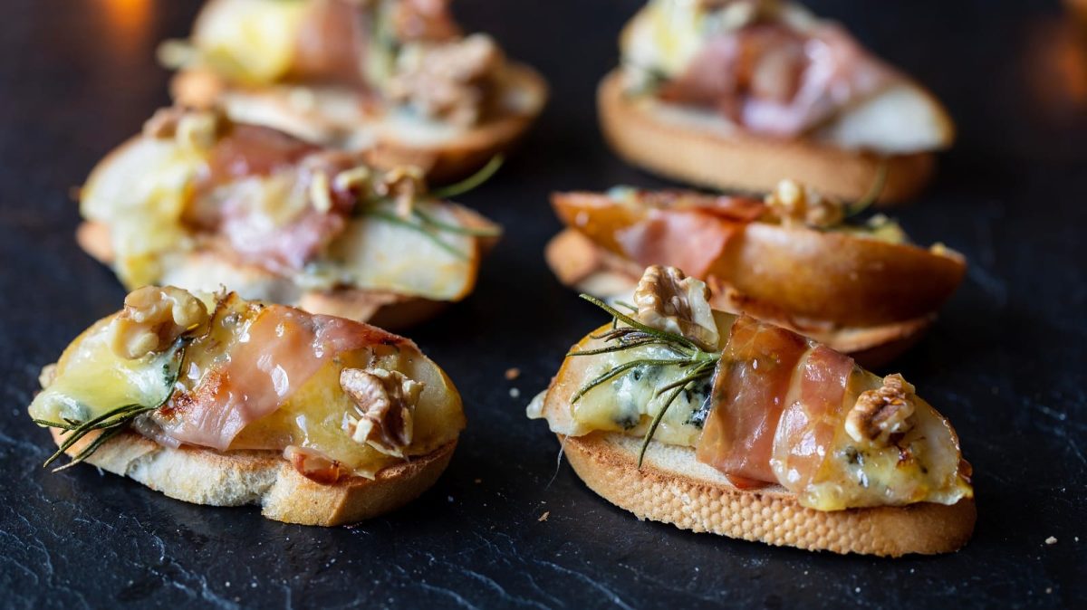 Eine Reihe Birnen-Canapés mit Gorgonzola und Speck auf einem schwarzen Tablett.