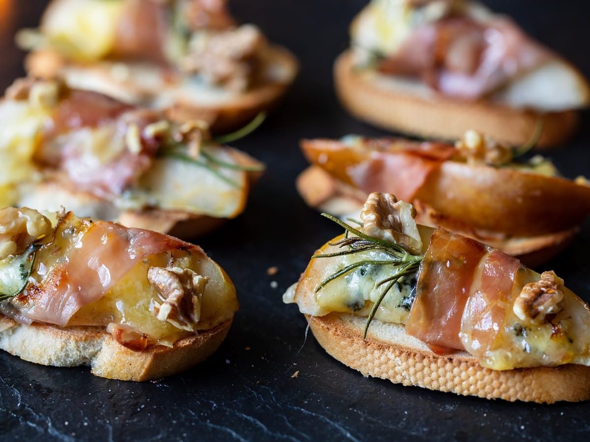 Eine Reihe Birnen-Canapés mit Gorgonzola und Speck auf einem schwarzen Tablett.