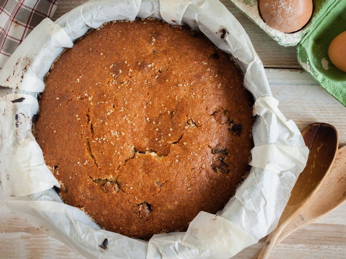 Draufsicht: Auf hellem Holz steht ein britischer Glühwein-Kuchen in einer runden Form. Drum herum liegen Eier und zwei Holzlöffel.