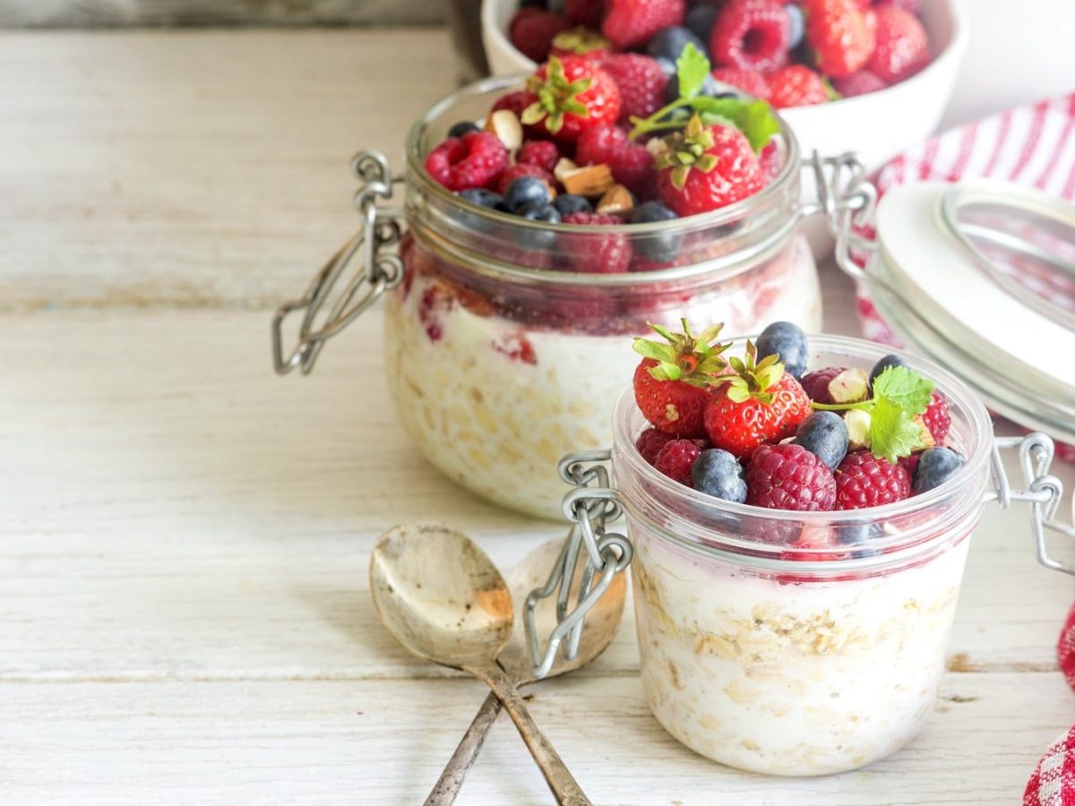 Zwei Weckgläser mit der Cheesecake Bowl und einer Menge Früchten auf einem hellen Holztisch von der Seite fotografiert. Daneben rot-weiß-karierte Tischdecken.