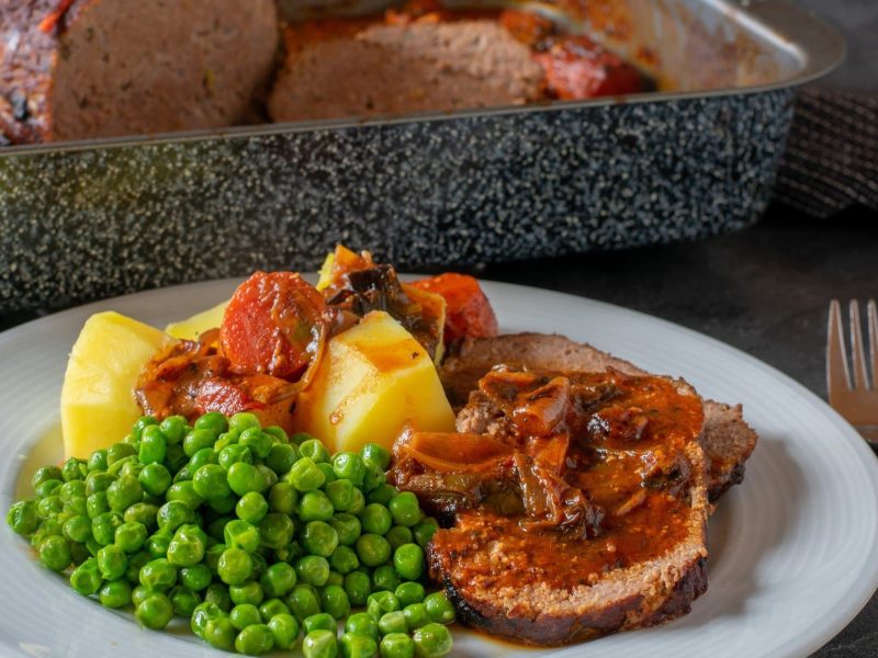 Draufsicht, schräg: Ein weißer Teller mit Omas Hackbraten mit Sauce und Erbsen steht auf einem Holztisch. Im Hintergrund ist der Hackbraten in einer Kastenform.