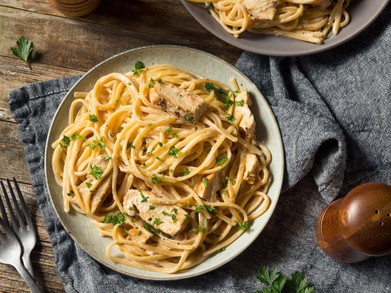 Draufblick: Zwei Teller mit Fettuccine mit Hühnchen auf einem Untergrund aus Holz und einem blauen Tuch. Daneben steht eine Pfeffermühle.