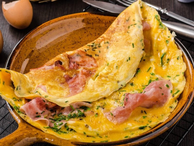 Draufsicht, schräg: Ein italienische Frittata al Prosciuttoin einer Pfanne auf gebranntem und glasiertem Ton. Daneben liegen Besteck und zwei aufgeschlagene Eier.