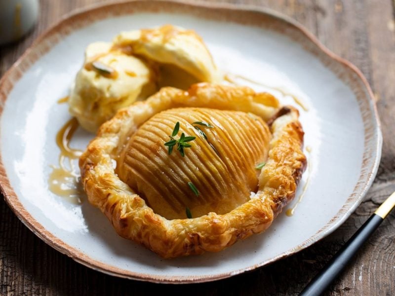 Die Hasselback-Birne in Blätterteig gebacken auf einem grauen Teller mit Eiscreme. Alles von schräg oben auf einem dunklen Holztisch fotografiert.