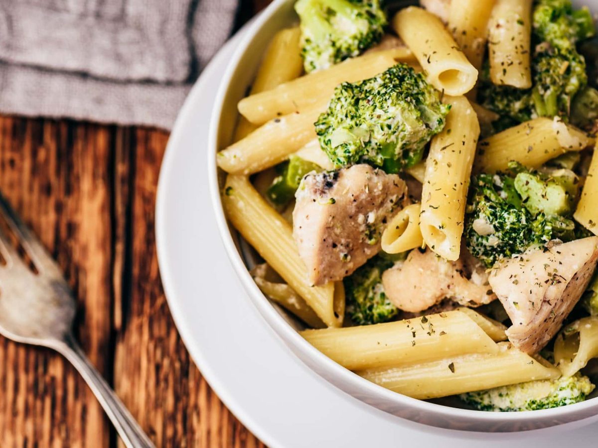 Draufsicht: Eine Schüssel Hühnchen-Pasta, die auf einem Teller steht. Daneben liegen eine Gabel und ein graues Tuch.