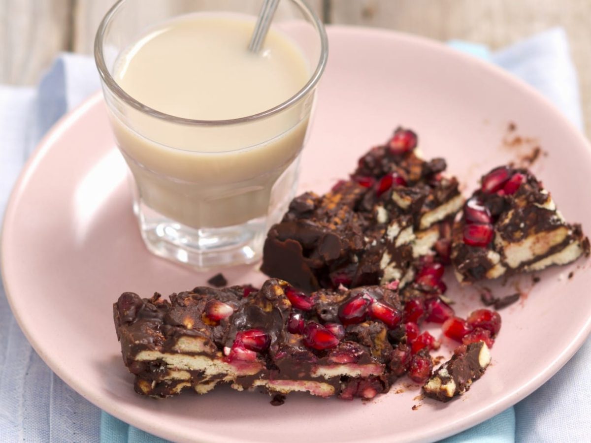 Eine Portion Kalter Hund-Kuchen mit Granatapfelkernen und ein Glas Milch auf einem rosa Teller. Der Teller steht auf einem hellblauen Geschirrtuch und der Hintergrund ist hölzern.