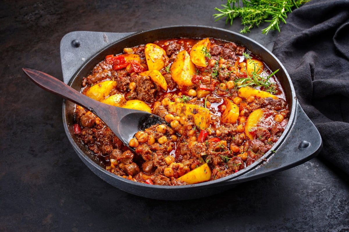 Draufsicht, schräg: eine Italienische Hackfleisch-Pfanne mit Kartoffeln steht auf einem schwarzen Untergrund. In der Pfanne ist ein Holzlöffel. Im Hintergrund ist frischer Rosmarin.