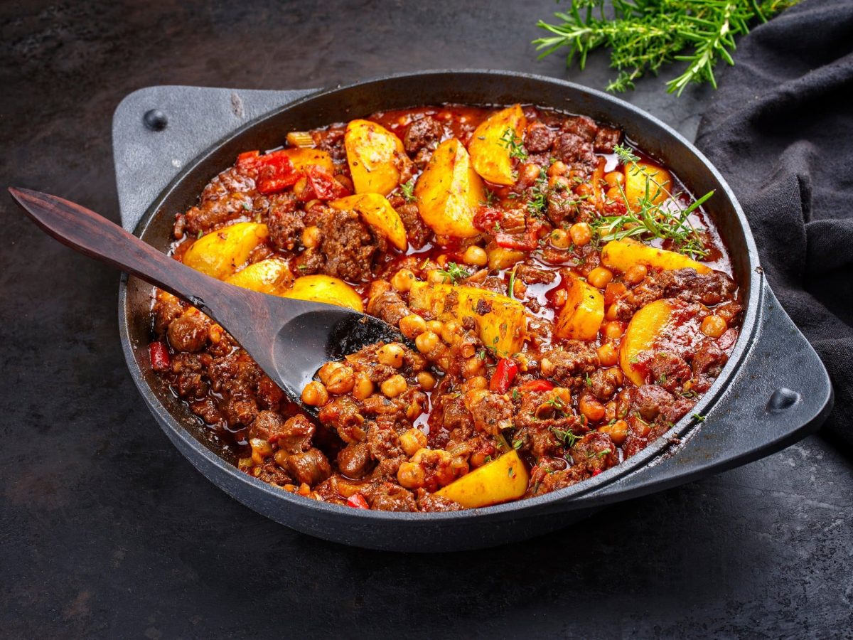 Draufsicht, schräg: eine Italienische Hackfleisch-Pfanne mit Kartoffeln steht auf einem schwarzen Untergrund. In der Pfanne ist ein Holzlöffel. Im Hintergrund ist frischer Rosmarin.