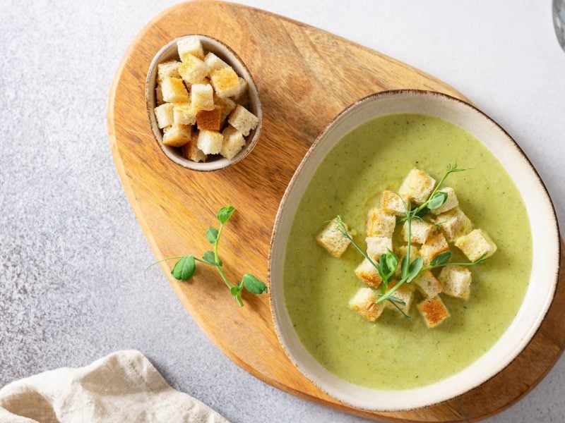 Eine weiße Schale mit der Suppe auf einem Holzbrett und weißem Steintisch. Alles direkt von oben fotografiert.