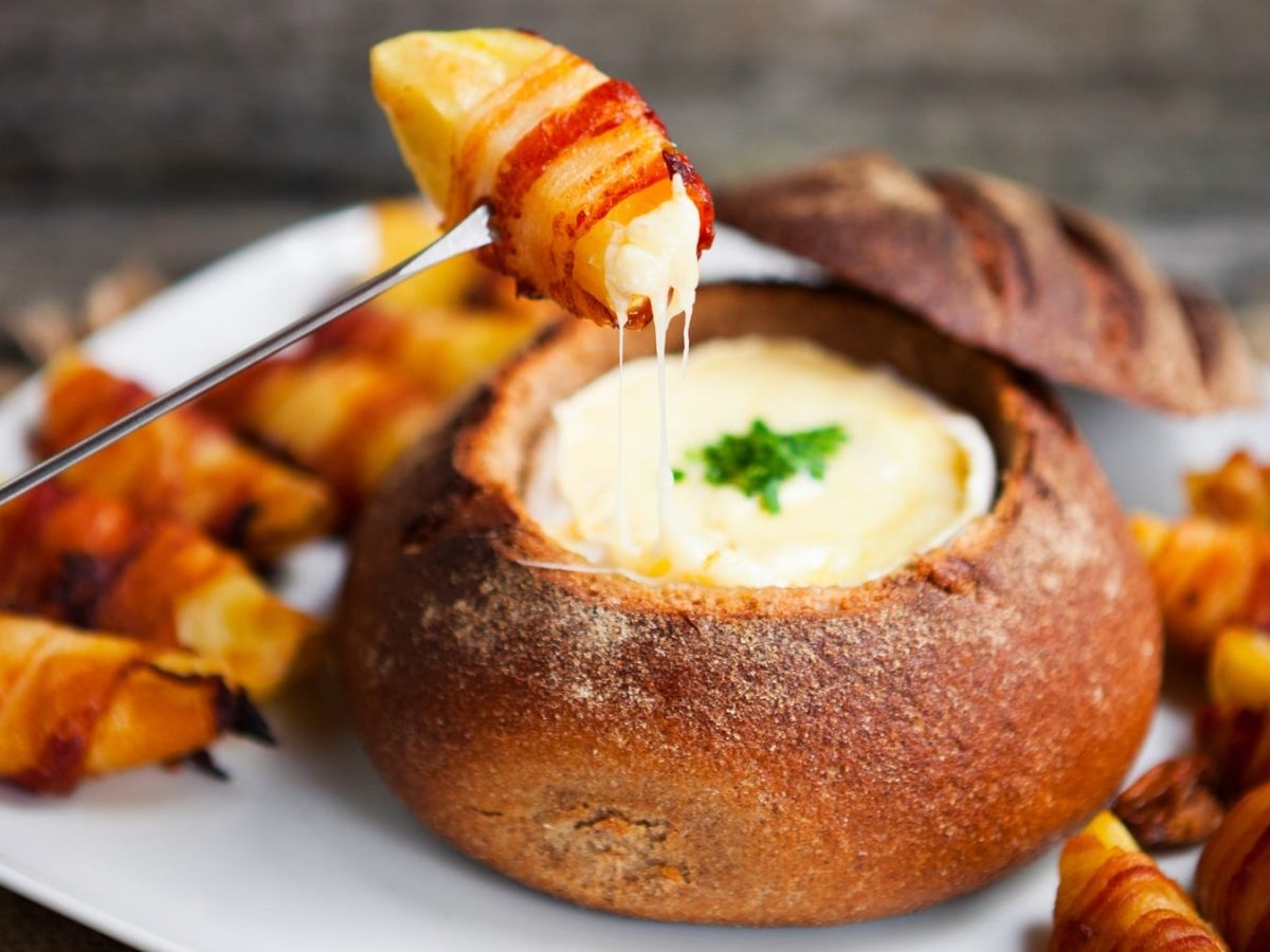 Das Fondue in einem ausgehöhlten Brotlaib mit den Speck-Kartoffeln auf einem weißen Teller und dunklem unter- und Hintergrund. Alles von leicht schräg oben fotografiert.