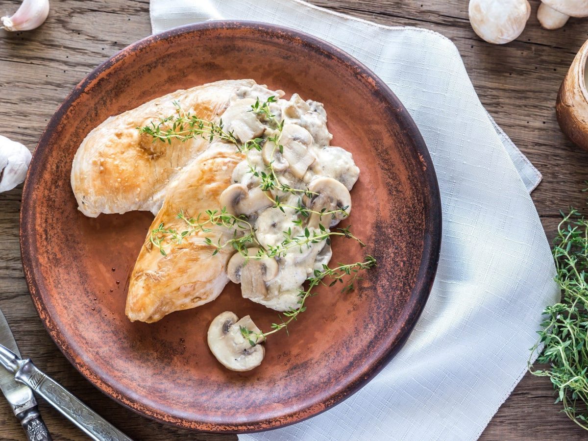 Draufsicht: Ein brauner Teller mit Knoblauch-Hühnchen in Sahne-Sauce liegt auf einem weißen Tuch, dass auf einer Holzplatte liegt. Drum herum sind ein Besteck und frischer Thymian.