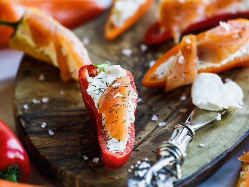 Draufsicht, schräg: Lachs-Häppchen in einer halbierten roten Spitzpaprika auf einem Holzbrettchen. Daneben liegt ein silbernen Messer.