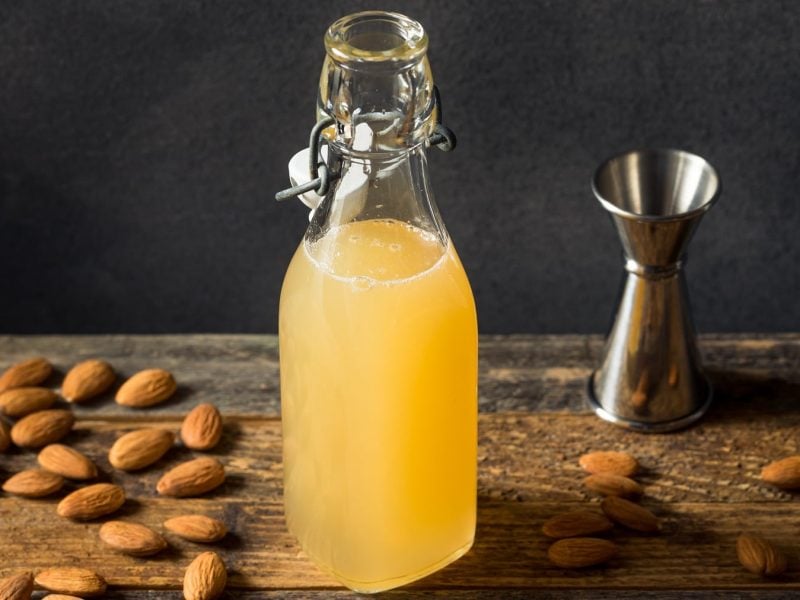 Eine Flasche mit dem Mandelsirup auf einem dunklen Holztisch mit Mandeln und einem Barmaß. Alles von der Seite fotografiert vor schwarzem Hintergrund.