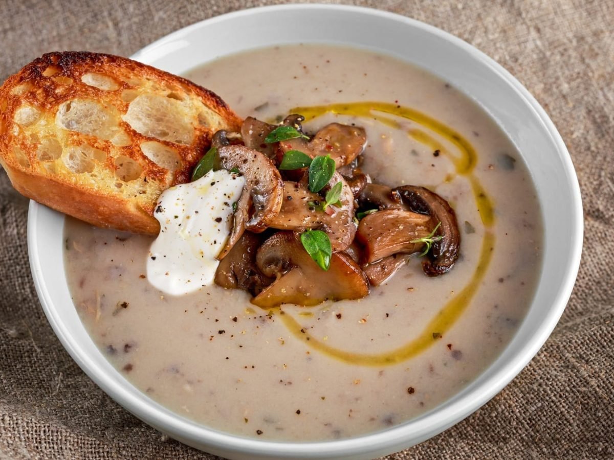 Eine weiße Schüssel mit der Maronen-Pilzcremesuppe auf einem beigen Leinentuch. In der Mitte der Suppe Pilze, Crème fraîche und geröstetes Brot.