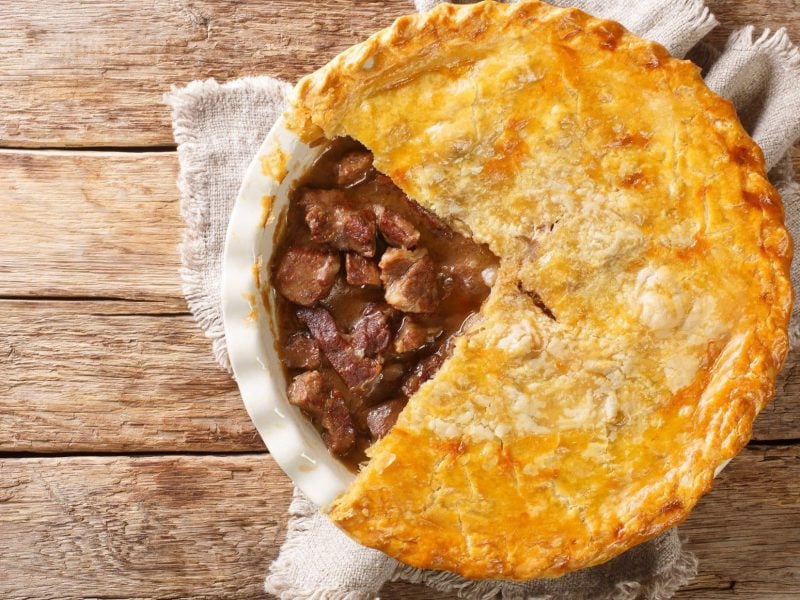 Draufsicht: Auf hellem Holz liegt eine weiße Auflaufform mit einem Meat Pie. Es handelt sich um einen Fleischkuchen. Der Meat Pie ist angeschnitten. Man sieht das gebratene Rindfleisch unter der Teighülle.