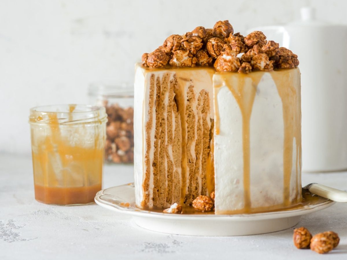 Ein Nusskuchen mit Karamell auf einem weißen Servierteller auf einem weißen Hintergrund. Links davon ein Glas mit Karamellsauce und dahinter eine Karaffe aus weißem Porzellan.