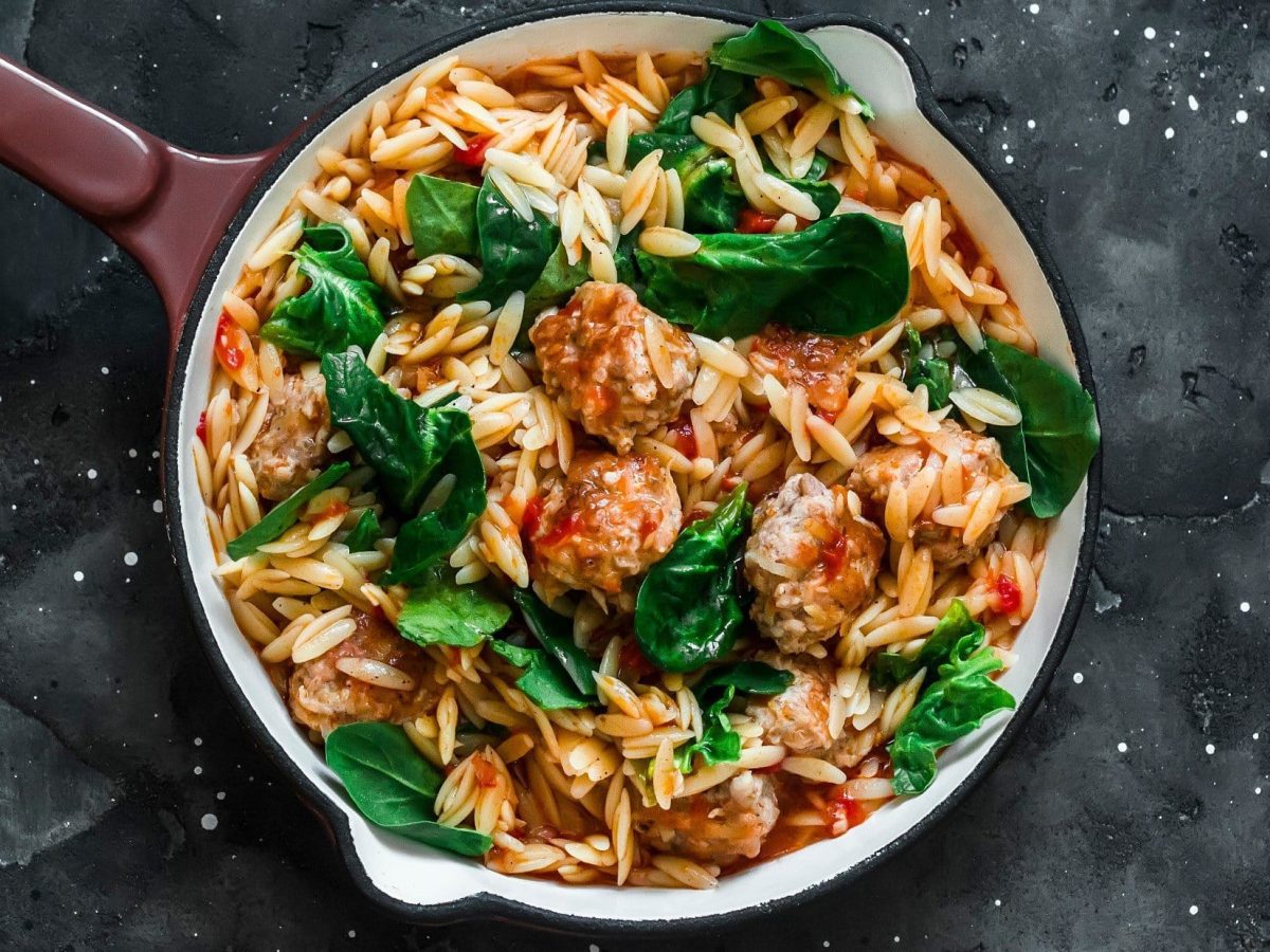 Draufblick: Ein Topf Orzo-Pasta mit Fleischbällchen auf dunklem Untergrund.