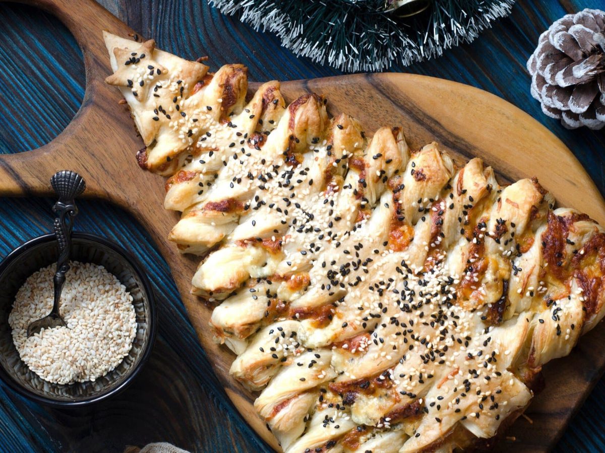 Draufsicht: Auf blauem Untergrund und einem Holzbrettchen liegt ein Puff Pastry Christmas Tree. Das ist ein Weihnachtssbaum aus Blätterteig.