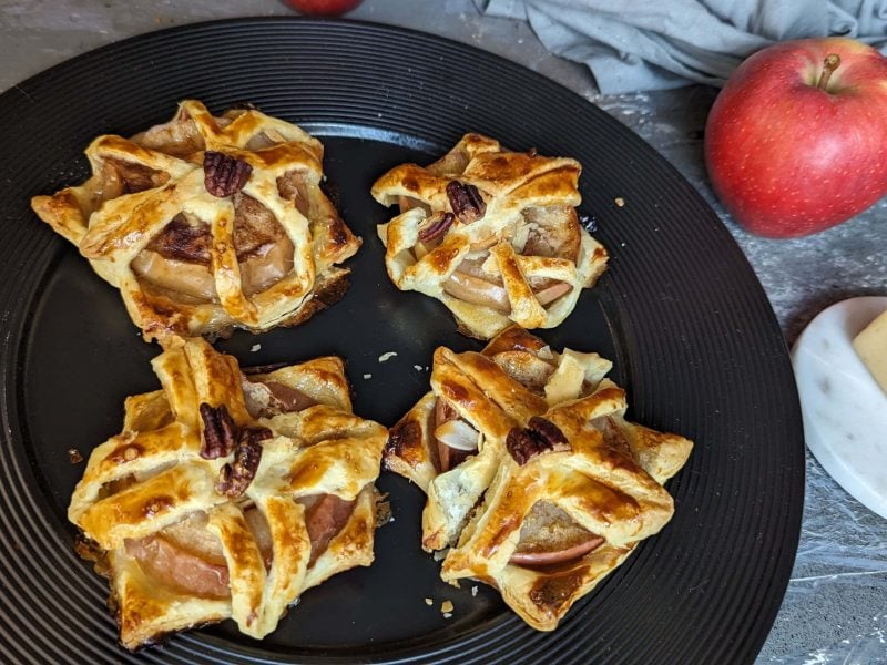 Vier Stücke Apfel-Marzipan-Plunder auf einem schwarzen Teller. Daneben Marzipan und ein roter Apfel. Alles auf einem grauen Tisch von schräg oben fotografiert.