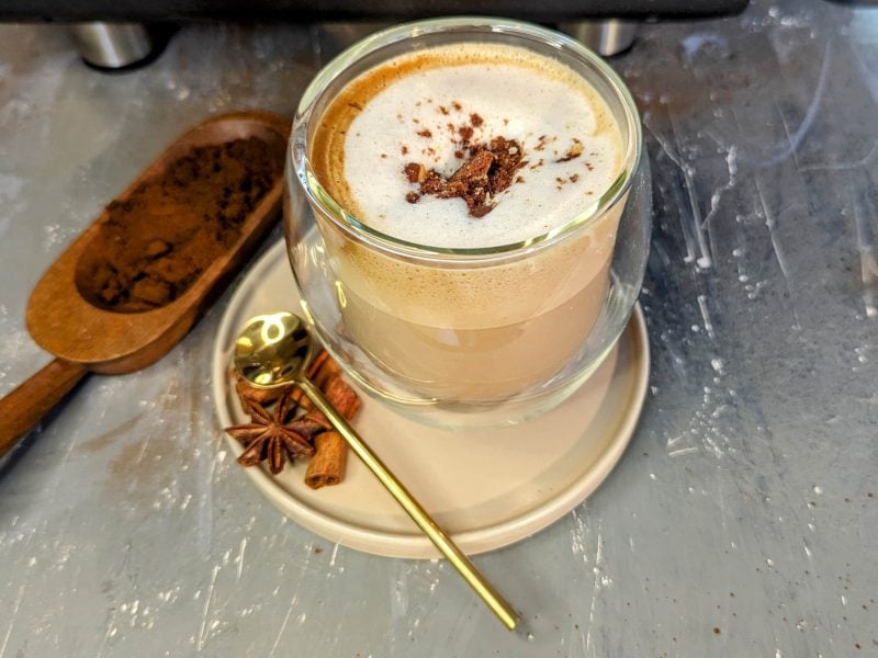 Ein Glas mit dem Kokos-Lebkuchen-Latte auf einem grauen Steintisch mit winterlichen Gewürzen und Kaffeepulver.