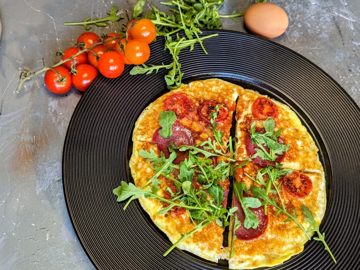 Ein schwarzer telle rmit der Frühstückspizza auf grauemj Untegrrund. Daneben Kirschtomaten, Ei und Rucola. Alles direkt von oben fotografiert.