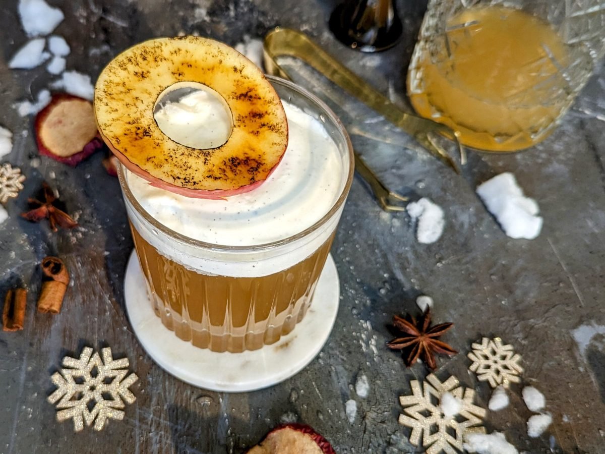 Ein Glas mit dem Old Fashioned Bratapfel auf grauem Untergrund. Daneben weihnachtliche Deko.